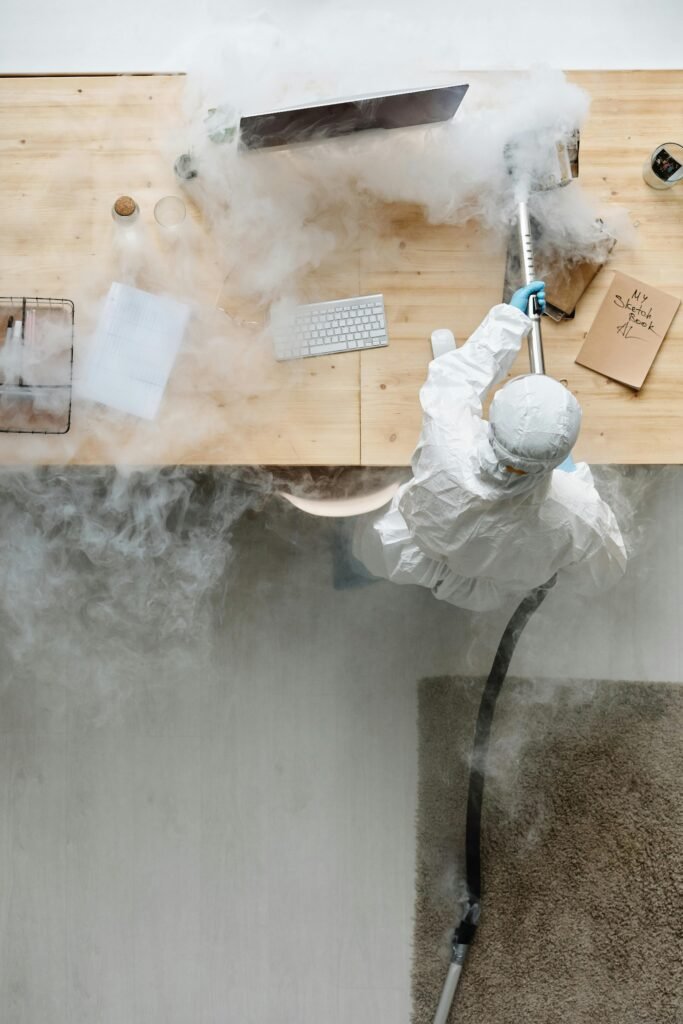 A person in protective gear sanitizing an office desk using fumigation equipment during daytime.